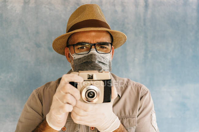 Homme d'âge moyen en gants de latex et masque médical prenant des photos avec caméra rétro en studio de lumière en regardant la caméra — Photo de stock