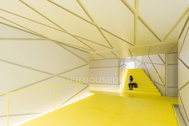 Feminino em desgaste casual sentado no corredor de escadas amarelo futurista com paredes geométricas e teto — Fotografia de Stock