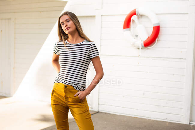 Calm female wearing trendy striped t shirt standing in port on background of wooden wall with lifesaver and looking at camera — Stock Photo