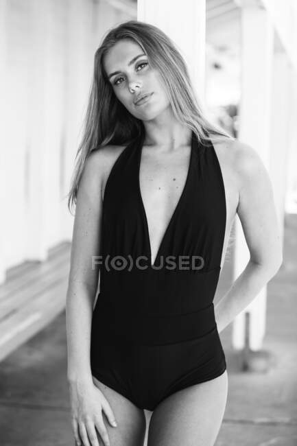 Black and white charming female in trendy swimwear leaning on column in port during holiday and looking at camera — Stock Photo