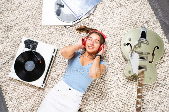 Сверху счастливая молодая женщина смотрит в повседневной одежде лежа на полу рядом с гитарой и слушать музыку с наушниками и виниловым проигрывателем, наслаждаясь свободное время дома — стоковое фото