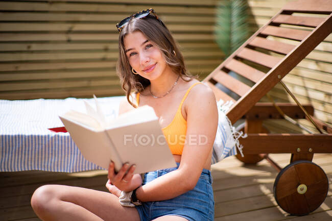 Веселая женщина в летнем топе и в шортах сидит возле шезлонга на деревянной веранде и наслаждается историей, улыбаясь и глядя в камеру — стоковое фото