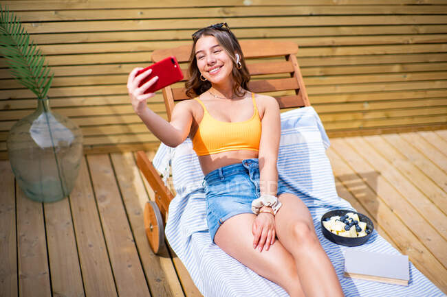 From above positive female in shorts and bra lying on deck chair on sunny day and taking photo on camera of cellphone — Stock Photo