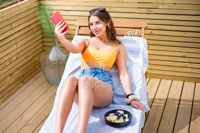 Зверху позитивна жінка в шортах і бюстгальтер лежить на стільці на сонячний день і фотографується на камеру мобільного телефону — стокове фото