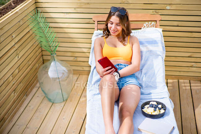 De cima positivo fêmea em shorts e sutiã deitado na cadeira de praia no dia ensolarado usando telefone celular — Fotografia de Stock