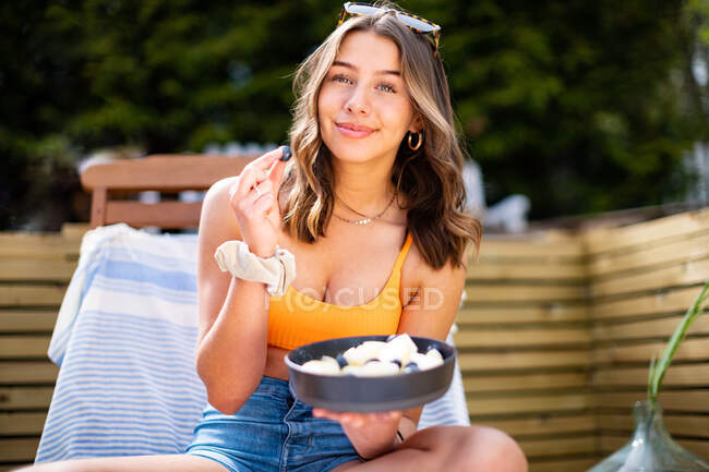 Mujer alegre en verano llevar sentado en la silla de cubierta con frutas maduras y bayas mientras se enfría en la terraza de madera y mirando a la cámara - foto de stock