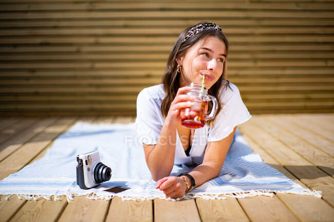 Весела молода жінка в повсякденному одязі п'є свіжий фруктовий коктейль і насолоджується сонячним літнім днем, лежачи на терасі біля миттєвої фотокамери — стокове фото