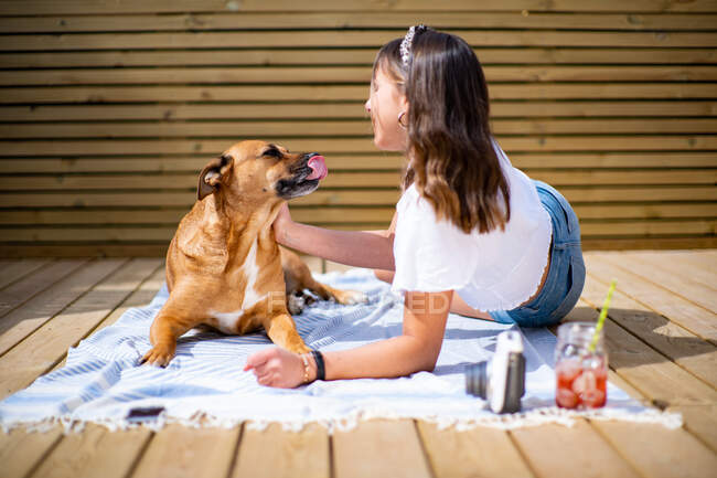 Side view of positive young female in casual shirt and jeans shorts sitting with cute dog lying nearby white resting on sunny terrace in summer day — Stock Photo