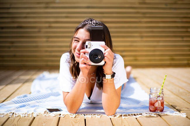 Позитивна молода жінка в повсякденній сорочці і джинсових шортах фотографує миттєву камеру, що лежить на сонячній терасі в літній день — стокове фото