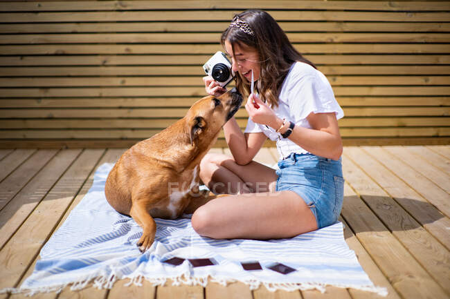 Vue latérale de la jeune femme positive en chemise décontractée et short en jeans prenant des photos avec caméra instantanée de chien mignon couché à proximité blanc reposant sur une terrasse ensoleillée dans la journée d'été — Photo de stock