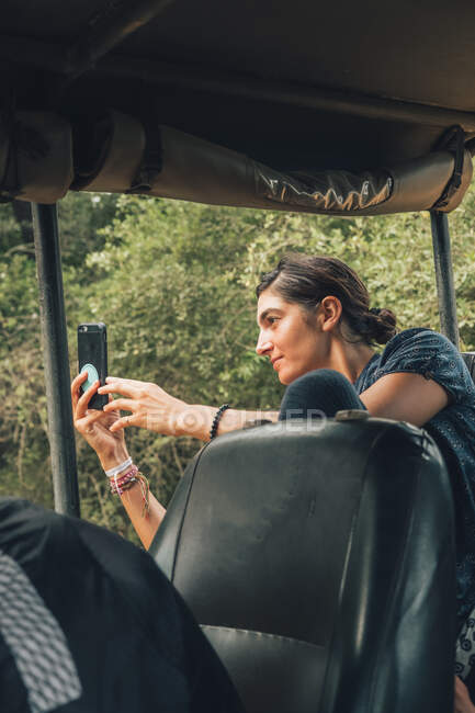 Side view of smiling female tourist sitting in car and taking pictures on smartphone during safari — Stock Photo