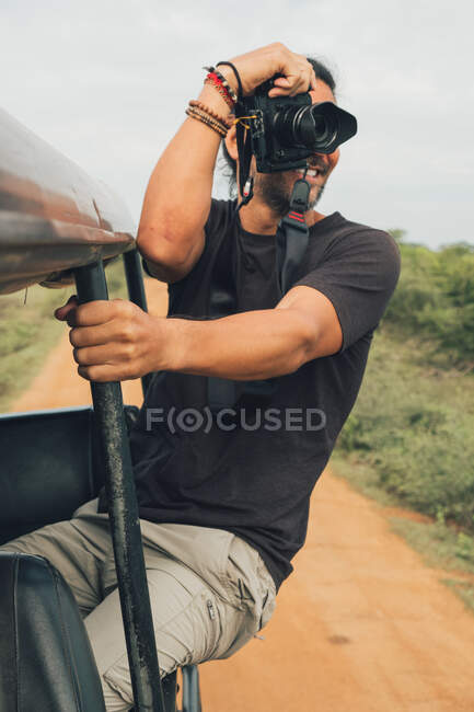 Fotógrafo masculino alegre étnico sentado no carro e tirando fotos da natureza durante as férias no safári — Fotografia de Stock