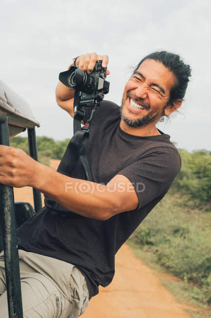 Fotógrafo masculino alegre étnico sentado en el coche y tomando fotos de la naturaleza durante las vacaciones en safari - foto de stock