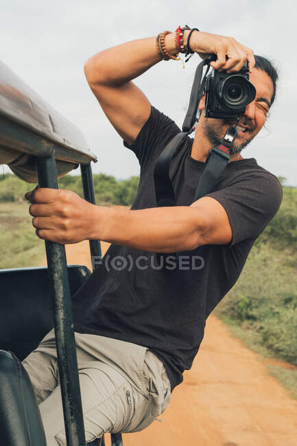 Fotógrafo masculino alegre étnico sentado no carro e tirando fotos da natureza durante as férias no safári — Fotografia de Stock