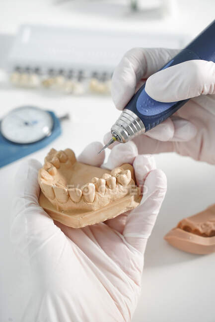 Сверху анонимный стоматолог в медицинских перчатках, шлифующий зубной протез профессиональным инструментом во время работы в современной лаборатории — стоковое фото