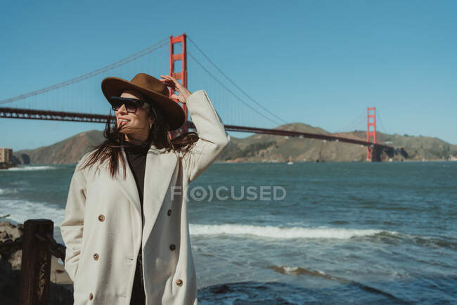 Seitenansicht einer lächelnden jungen Frau im trendigen Outfit mit Hut und Sonnenbrille, die bei sonnigem Wetter auf einer Böschung vor der Golden Gate Bridge in Kalifornien steht — Stockfoto