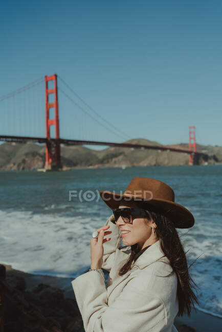 Vista lateral de sorrir jovem senhora na roupa da moda com chapéu e óculos de sol em pé no dique contra Golden Gate Bridge, na Califórnia, em dia ensolarado — Fotografia de Stock