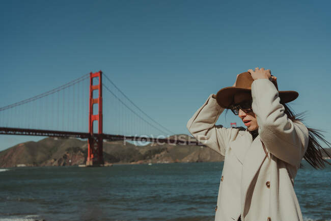 Seitenansicht einer jungen Frau im trendigen Outfit mit Hut und Sonnenbrille, die bei sonnigem Wetter auf einer Böschung vor der Golden Gate Bridge in Kalifornien steht — Stockfoto