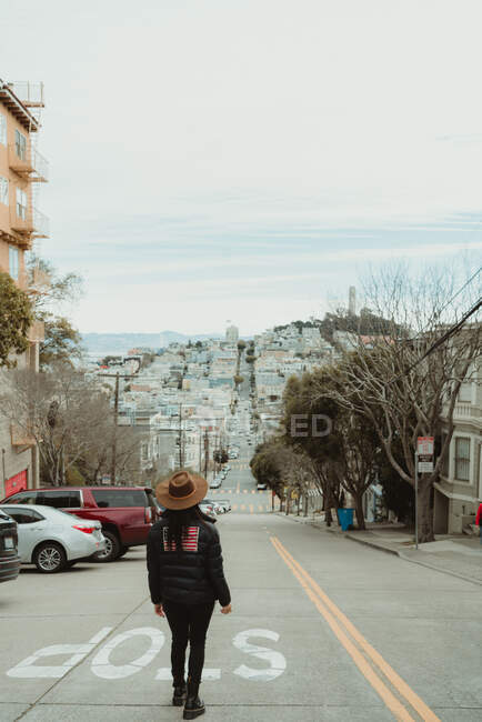 Back view of unrecognizable female traveler in trendy outfit and hat standing near stop sign on roadway leading towards hill during stroll and sightseeing in San Francisco city in cloudy day — Stock Photo