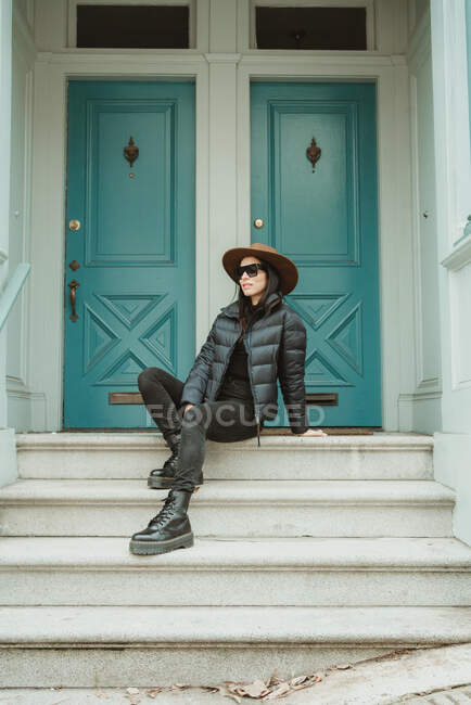 Low angle of confident young female dressed in trendy black jacket and jeans with hat and sunglasses sitting on stone stairs of old building with closed green doors on street of San Francisco city — Stock Photo