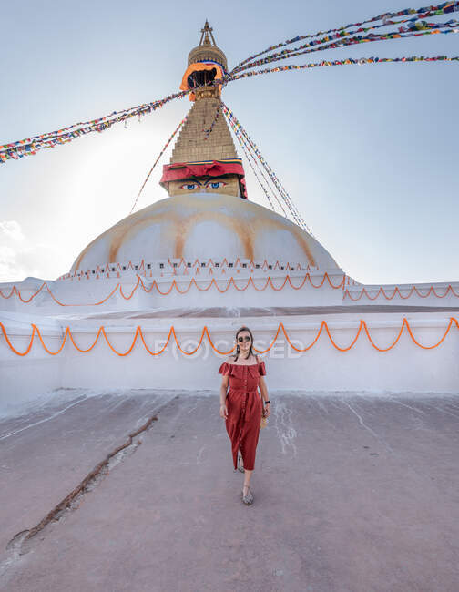 Mujer feliz mirando a la cámara de pie cerca del templo budista con guirnaldas decorativas y torre bajo el cielo nublado a la luz del día - foto de stock