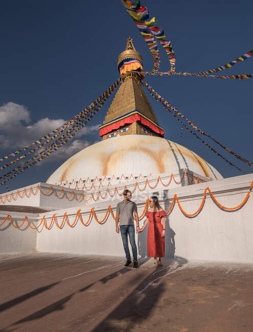 Paar hält Händchen und schaut einander an, während es in der Nähe des buddhistischen Tempels mit dekorativen Girlanden und Turm unter wolkenverhangenem Himmel im Tageslicht steht — Stockfoto