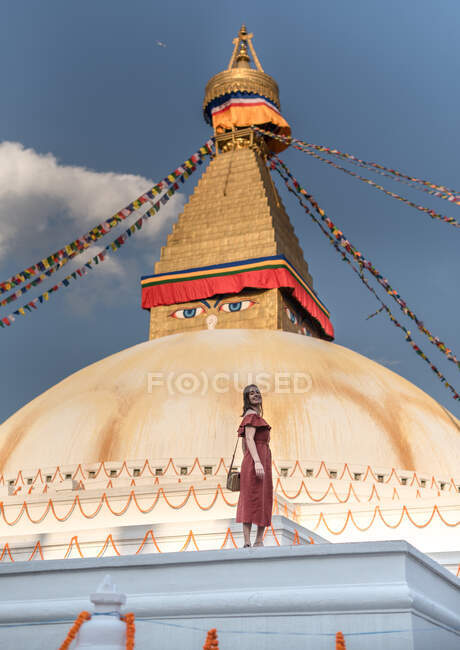 Donna felice guardando la macchina fotografica in piedi vicino al tempio buddista con ghirlande decorative e torre sotto cielo nuvoloso alla luce del giorno — Foto stock