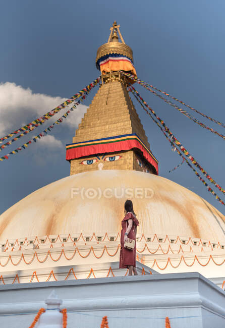 Vista laterale di donna irriconoscibile in piedi vicino al tempio buddista con ghirlande decorative e torre sotto il cielo nuvoloso alla luce del giorno — Foto stock