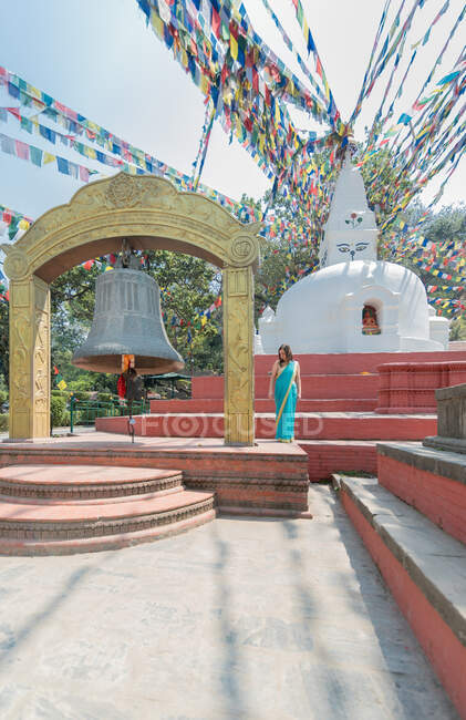 Touriste féminine en robe regardant loin tout en se tenant sur le trottoir près de vieux stupa hémisphérique en pierre avec coupole sur le dessus sous guirlande avec drapeaux et grande cloche en été — Photo de stock