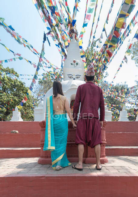 Back view of unrecognizable couple holding hands while standing on red stairs looking at ancient sculpture and arch with big bell under garland with prayer flags — Stock Photo