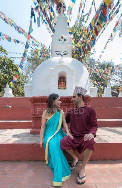 Couple holding hands and looking at each other while sitting on red stairs near ancient sculpture and arch under garland with prayer flags — Stock Photo