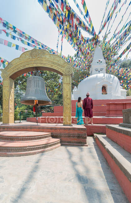 Back view of unrecognizable couple holding hands while standing on red stairs looking at ancient sculpture and arch with big bell under garland with prayer flags — Stock Photo