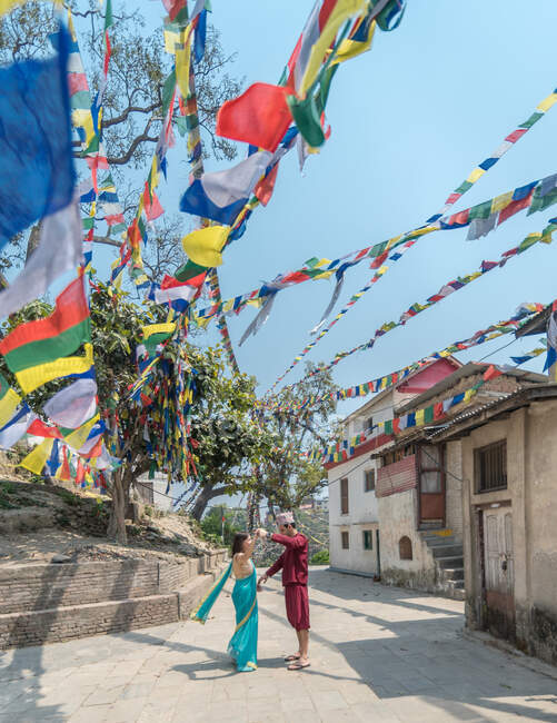 Vista lateral do casal em desgaste casual andando perto de edifícios budistas antigos sob guirlanda colorida com bandeiras no dia ensolarado — Fotografia de Stock