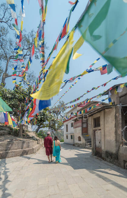 Visão traseira do casal anônimo em desgaste casual andando perto de edifícios budistas antigos sob guirlanda colorida com bandeiras no dia ensolarado — Fotografia de Stock