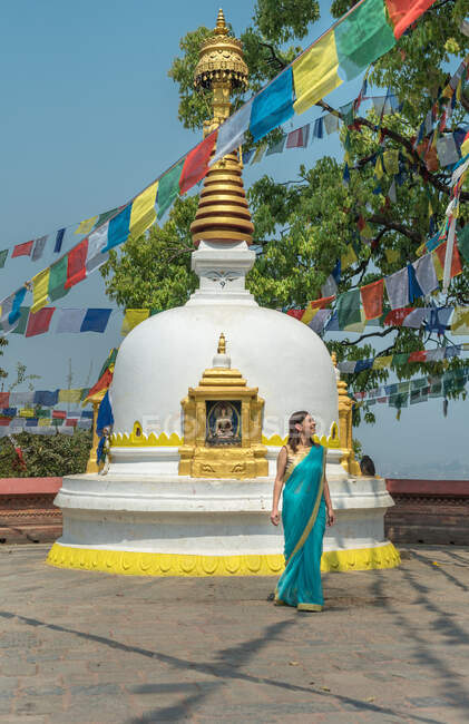 Turista feminina em vestido olhando para longe, enquanto em pé no pavimento perto de pedra velha stupa hemisférico com cúpula no topo sob guirlanda com bandeiras no verão — Fotografia de Stock