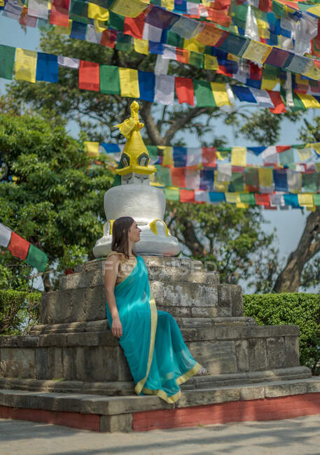 Touriste féminine en robe regardant loin tout en étant assis sur une vieille pierre sur un stupa bouddhiste avec coupole sur le dessus sous guirlande avec des drapeaux en été — Photo de stock