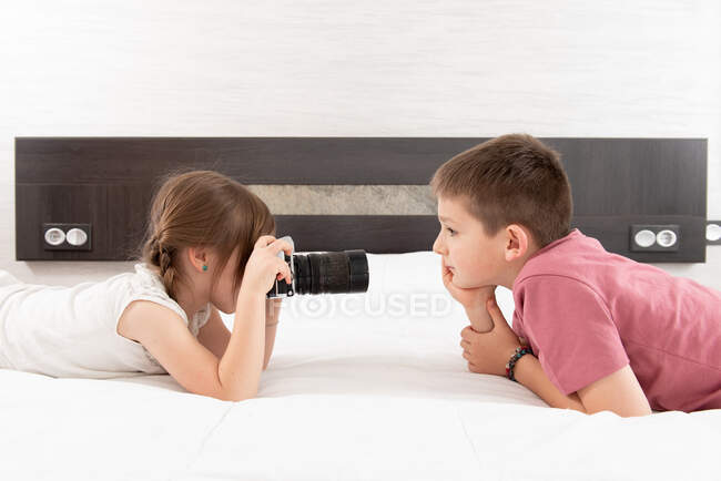 Side view of cute sister taking picture of brother on photo camera while lying on bed at home — Stock Photo