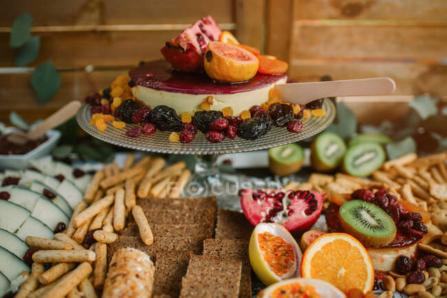 Вкусный чизкейк на подносе, украшенный различными фруктами подается на стол с разнообразной едой — стоковое фото
