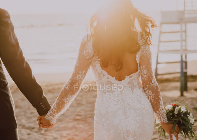 Visão traseira do noivo da colheita em terno elegante e noiva elegante no vestido de noiva e com buquê andando ao longo da praia durante o pôr do sol — Fotografia de Stock