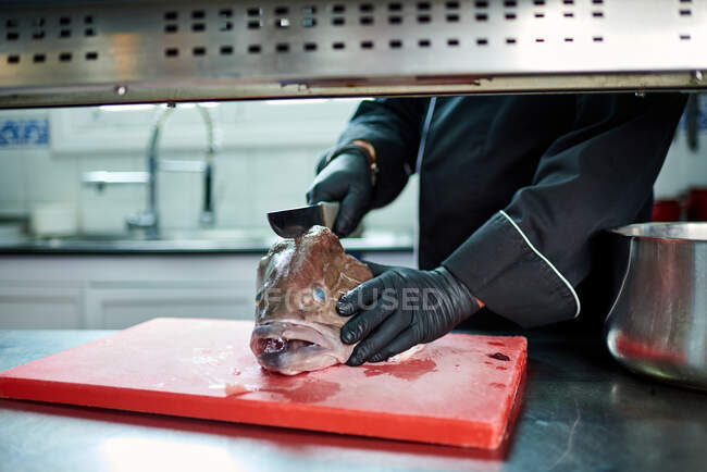 Cocinero irreconocible en uniforme negro y guantes usando una cuchilla de metal mientras corta la cabeza de un gran pescado fresco en una tabla picada roja en la mesa contra el interior borroso de la cocina contemporánea - foto de stock