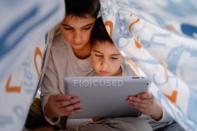 Siblings in pajamas hiding under blanket and enjoying interesting cartoon during daytime at home — Stock Photo