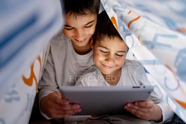 Hermanos positivos en pijama escondidos debajo de la manta y disfrutando de  dibujos animados interesantes durante el día en casa — Feliz, Adorable -  Stock Photo | #384274748