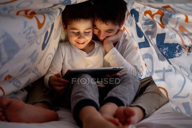Hermanos positivos en pijama escondidos debajo de la manta y disfrutando de dibujos animados interesantes durante el día en casa - foto de stock