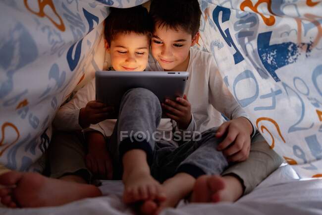 Positive Geschwister im Pyjama, die sich unter einer Decke verstecken und tagsüber zu Hause interessante Zeichentrickfilme genießen — Stockfoto