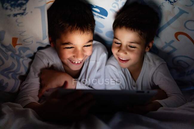 Frères et sœurs positifs en pyjama se cachant sous la couverture et appréciant un dessin animé intéressant pendant la journée à la maison — Photo de stock