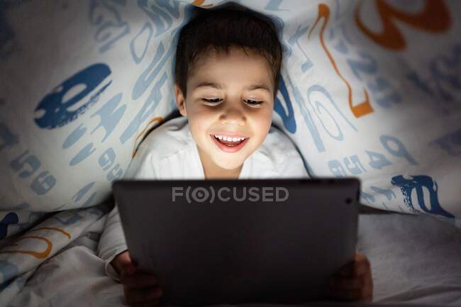 Очаровательный ребенок в пижаме прячется под одеялом и использует планшет во время развлечений в ночное время дома — стоковое фото