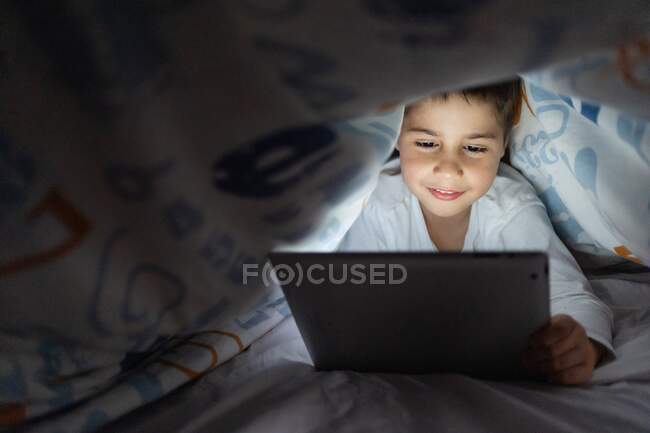 Adorable niño en pijama escondido debajo de la manta y el uso de la tableta mientras se entretiene durante la noche en casa - foto de stock