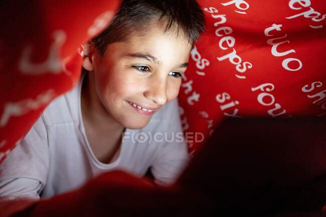 Criança adorável em pijama escondido sob cobertor e usando tablet enquanto se diverte durante a noite em casa — Fotografia de Stock