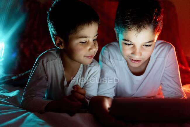 Hermanos positivos en pijama escondidos debajo de la manta y disfrutando de  dibujos animados interesantes durante el día en casa — Joy, acogedor -  Stock Photo | #384280868
