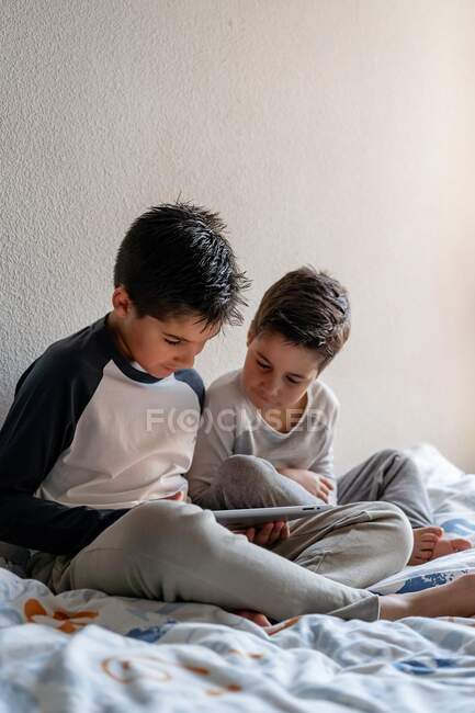 Hermanos en ropa de dormir sentados en una cama acogedora y viendo películas juntos mientras se entretienen en casa - foto de stock